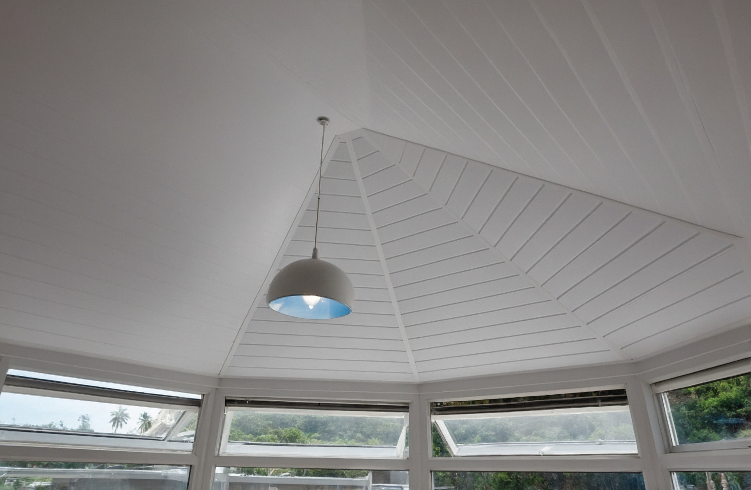 Interior of a conservatory showcasing a white, paneled warm roof and a modern hanging light fixture, with large windows offering a view of greenery outside.